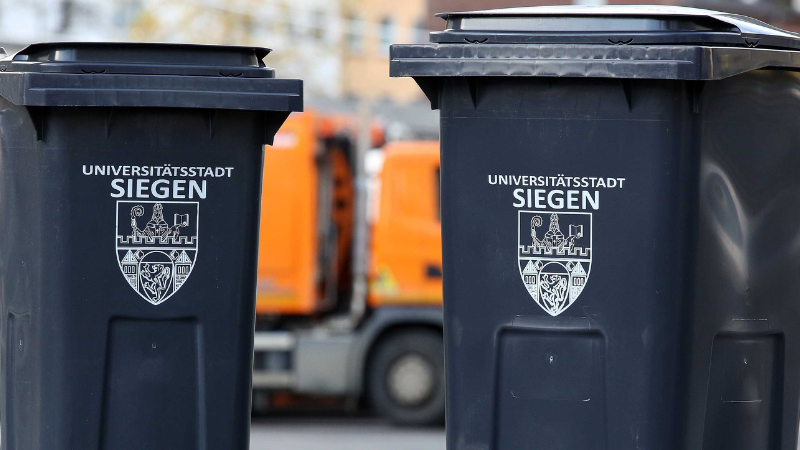 Symbolfoto: Müllabfuhr. Zu sehen sind zwei graue Tonnen mit dem Siegener Stadtwappen drauf. Im Hintergrund ist ein Müllfahrzeug zu erkennen. (Foto: Stadt Siegen)