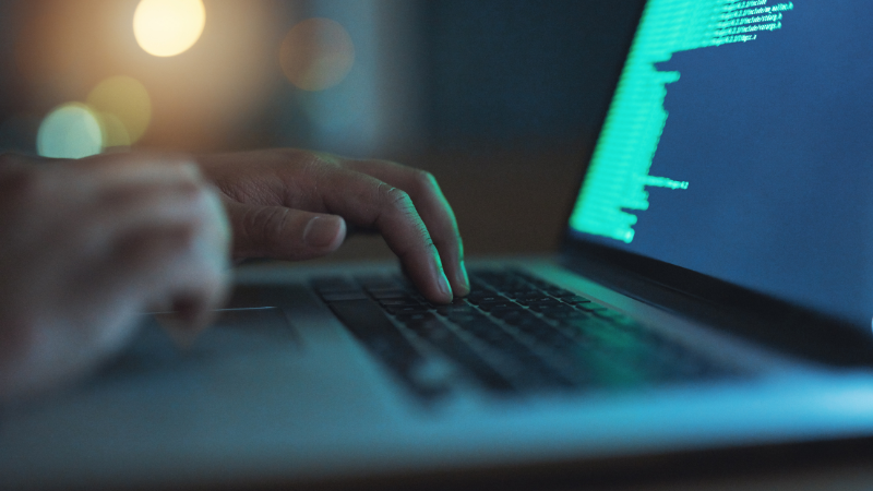 Symbolfoto: Cyber-Angriff. Zu sehen ist eine Nahaufnahme eines Laptops, auf dessen Bildschrim eine Reihe von Zahlen und Buchstaben zu erkennen ist. Rechts im Bild sind Hände abgebildet, die auf der Tastatur liegen.